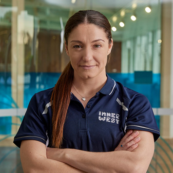 Personal Trainer Belinda wearing a navy blue shirt with an Inner West logo looking at the camera with arms crossed